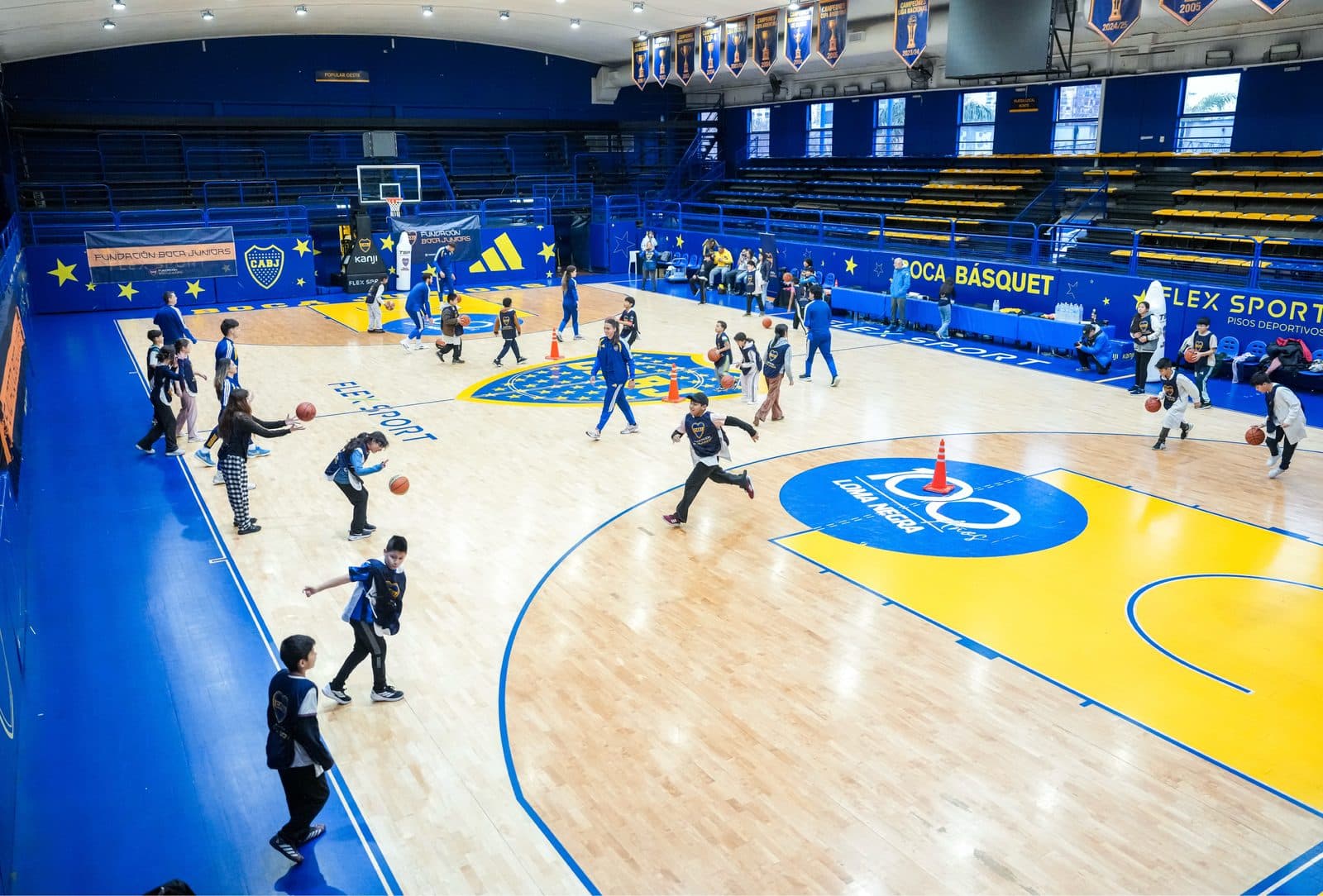 Chicos y chicas jugando al básquet en el parquet de La Bombonerita, con el escudo del club pintado en el piso y el cartel de Boca Básquet al fondo
