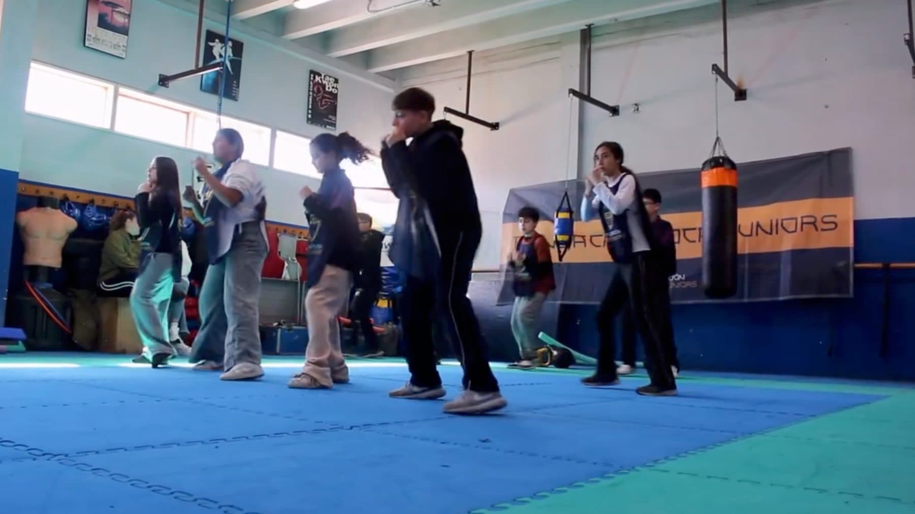 Chicos y chicas en posición de guardia sobre el tatami del gimnasio, con la bolsa de boxeo y el banner de la Fundación Boca Juniors al fondo