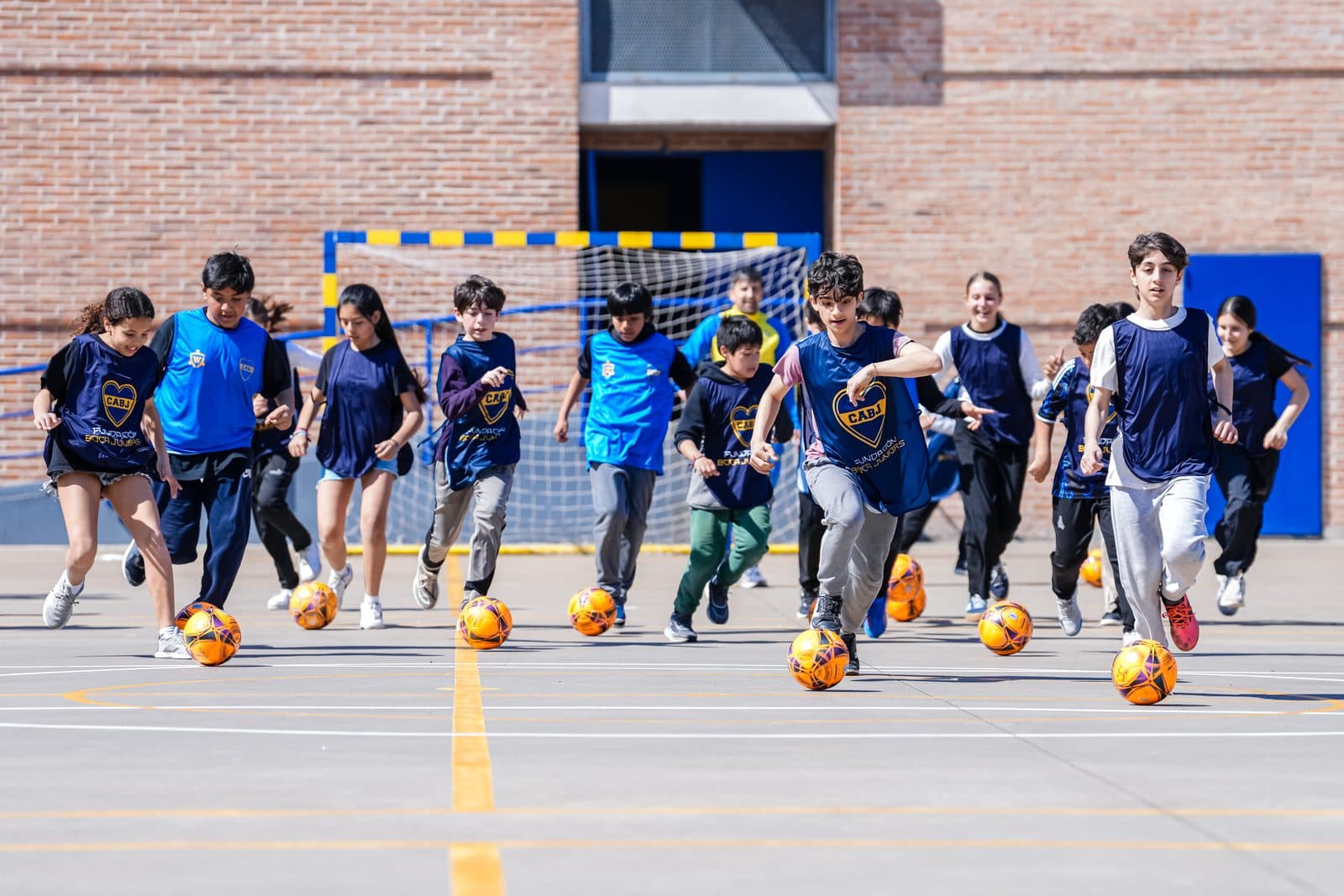 Chicos y chicas con pecheras de la Fundación Boca Juniors conducen pelotas de futsal en la cancha, con el arco y el paredón de ladrillo detrás