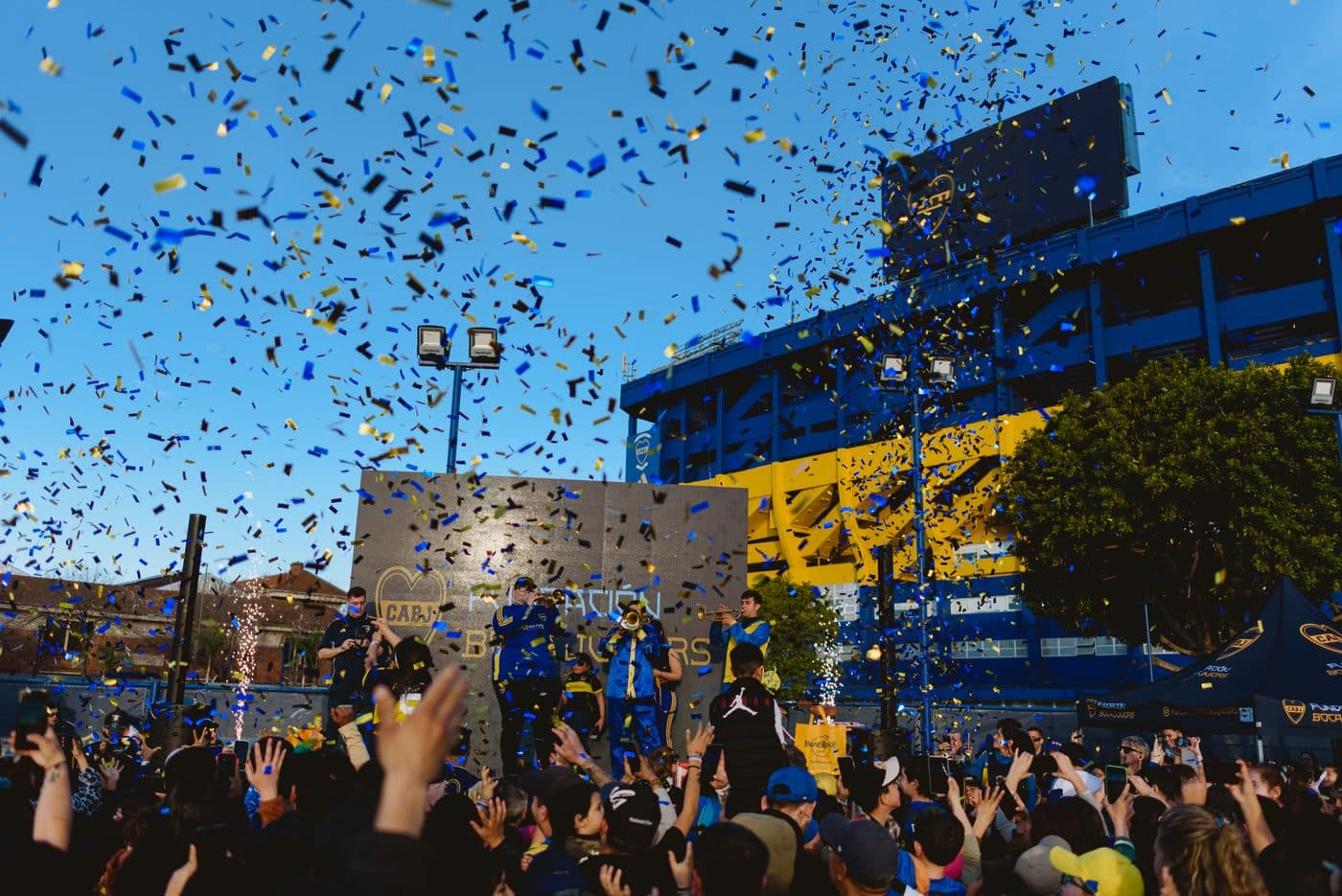 Papelitos azules y dorados cayendo sobre la multitud mientras la murga toca frente al muro de la Fundación Boca Juniors, con la Bombonera detrás
