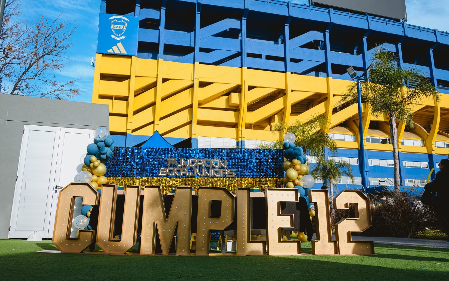 Letras luminosas gigantes que dicen CUMPLE 12 montadas en el césped, con la tribuna de la Bombonera de fondo