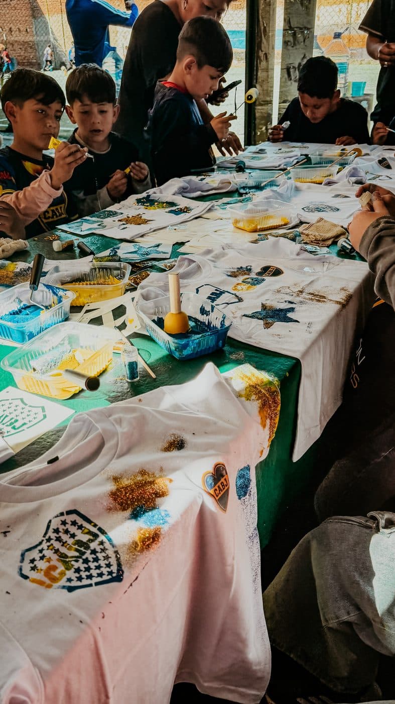 Las mesas del taller cubiertas de camisetas, pinceles y pinturas, con los chicos trabajando alrededor