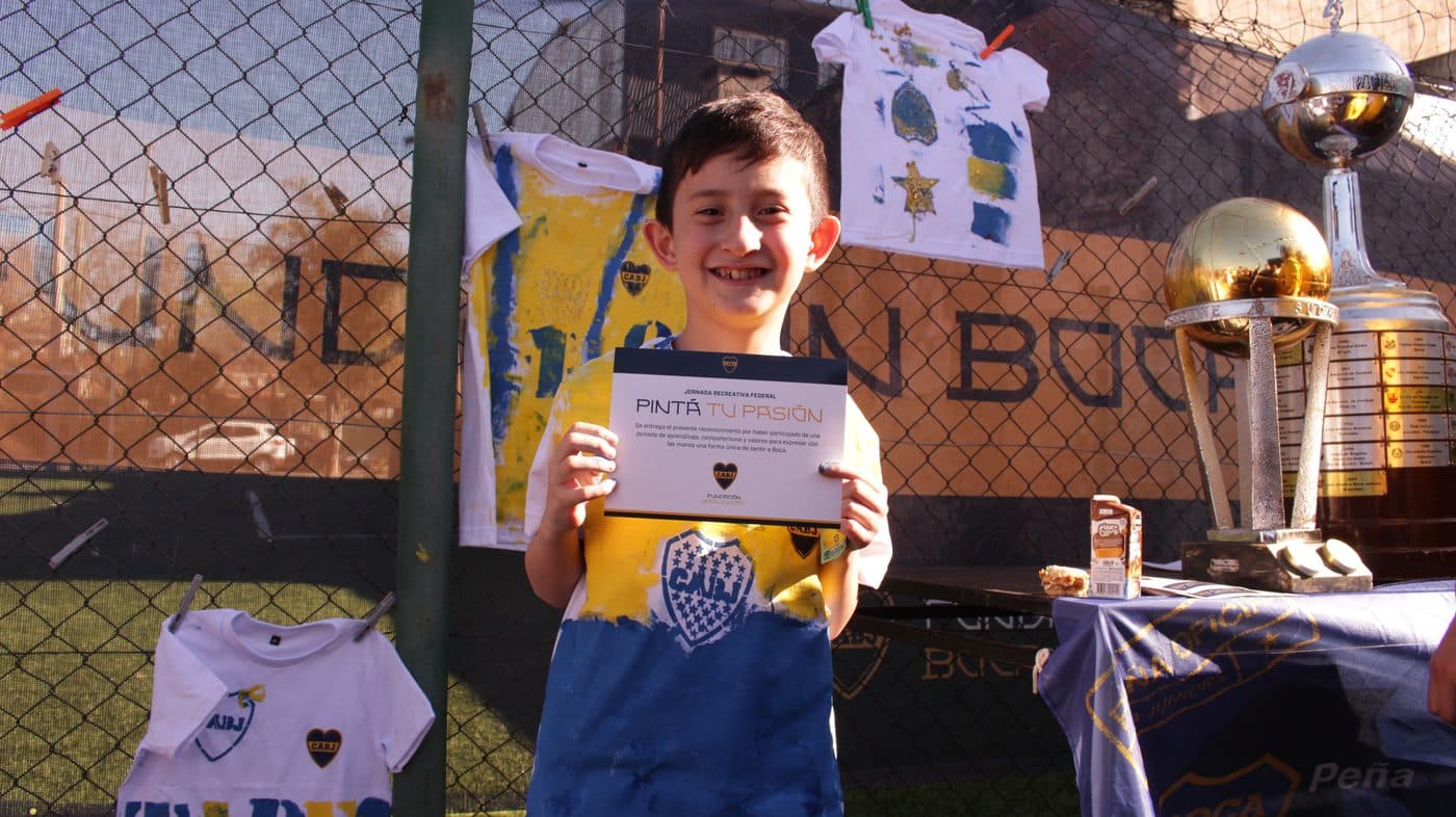 Un chico sonríe mostrando su certificado, con las camisetas pintadas colgadas detrás