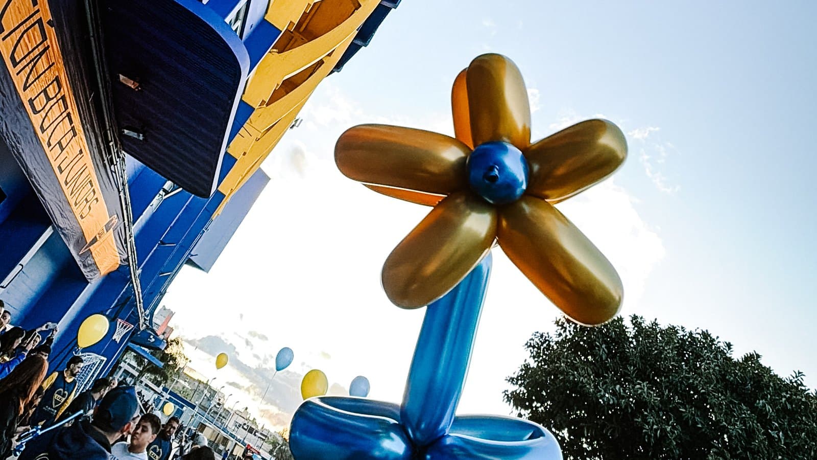 Una flor hecha con globos azules y dorados sostenida en alto frente al edificio de la Fundación, con las familias y los globos de la jornada abajo
