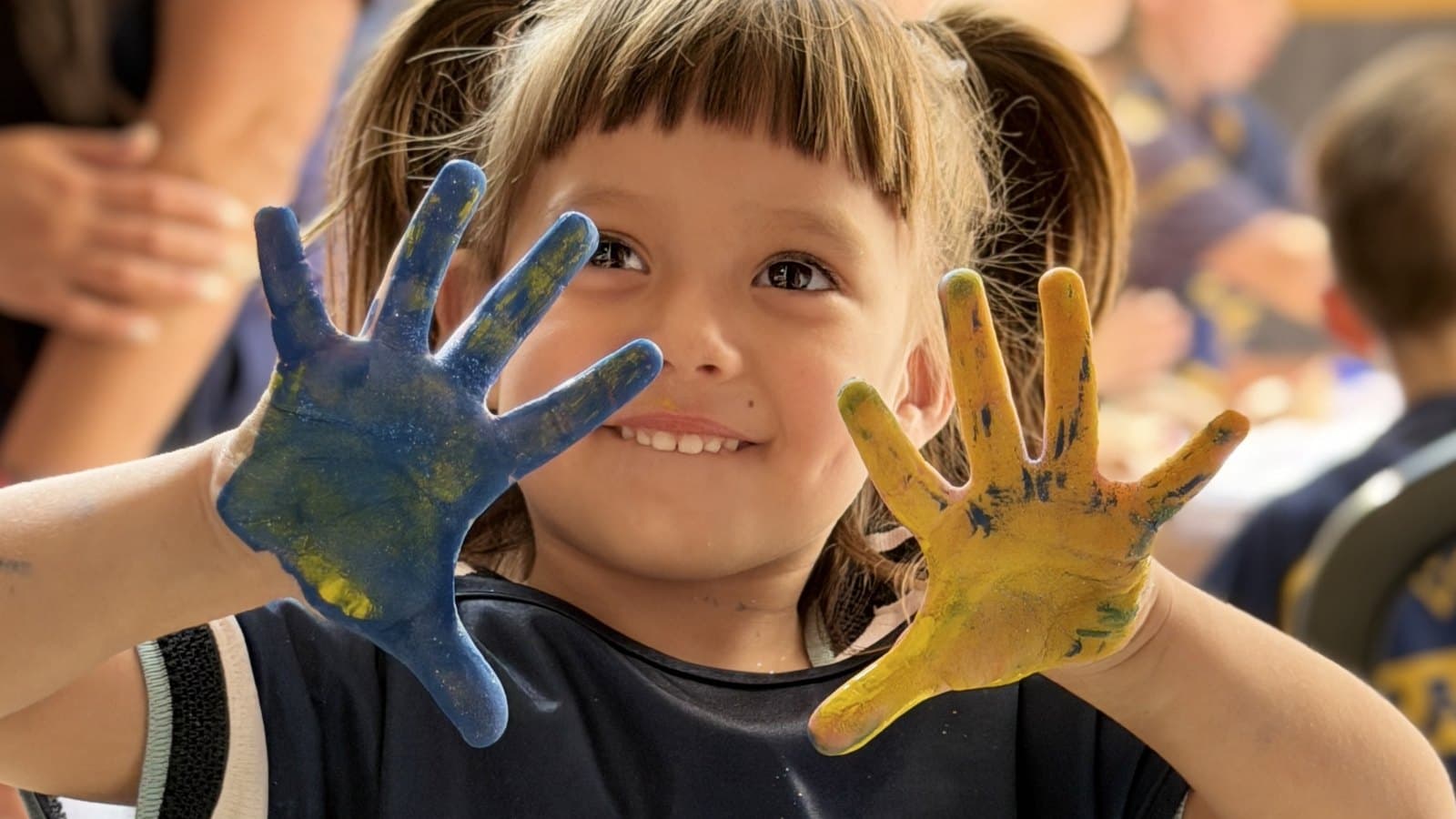 Una nena con la pechera de la Fundación muestra las manos pintadas de azul y de amarillo, sonriendo frente a la cámara