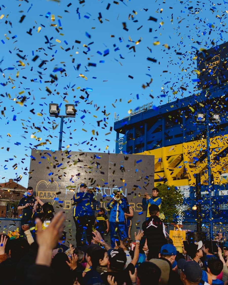 La murga tocando sobre el escenario entre papelitos azules y dorados, con la Bombonera y el muro de la Fundación detrás
