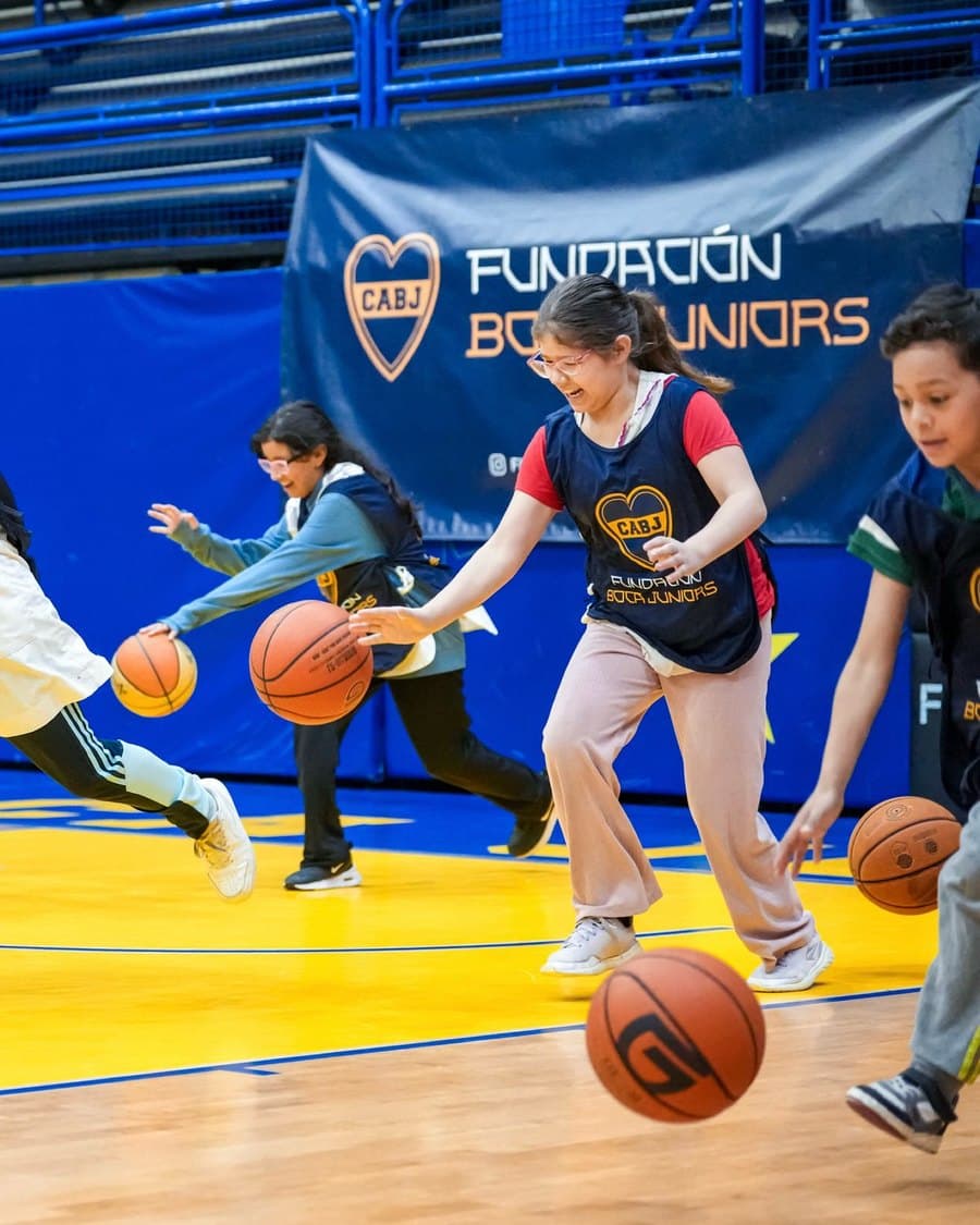 Chicas y chicos picando la pelota sobre el parquet de La Bombonerita, con el banner de la Fundación Boca Juniors detrás